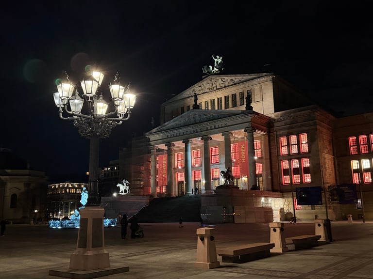 Historic theater building illuminated at night with red-lit facade, ornamental street lamp, and plaza seating in foreground