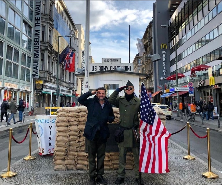 Two soldiers in uniforms posing with US flags in front of Checkpoint Charlie sign in Berlin